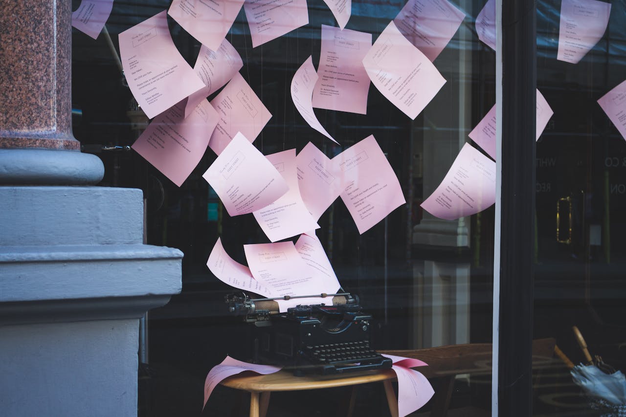 Artistic display of a vintage typewriter with pink papers floating in a window setting, indoors.